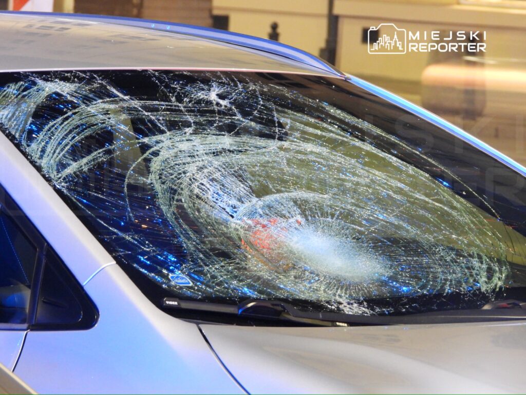 Szyba przednia samochodu z widocznymi pęknięciami i uszkodzeniami, w tle miejskie otoczenie.