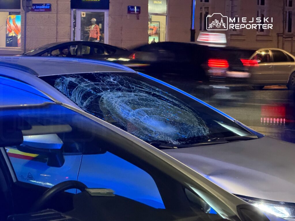 Uszkodzona szyba samochodowa z widocznymi śladami uszkodzenia, w tle nocne ulice oświetlone przez latarnie.