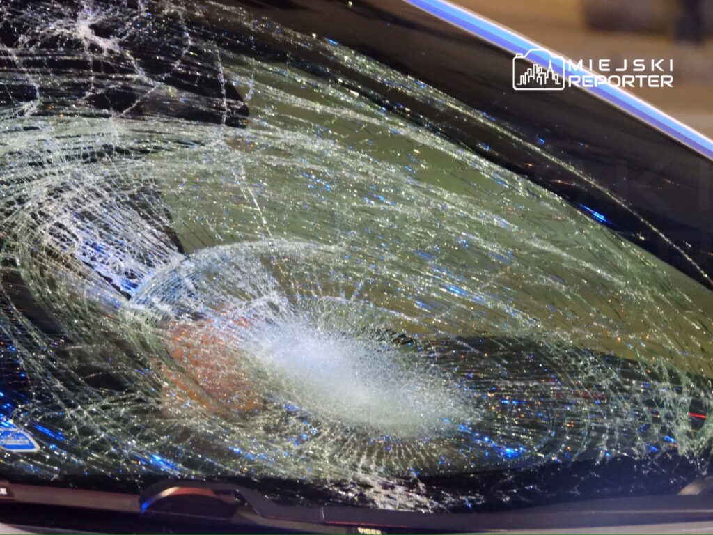 Zbita szyba samochodowa z wyraźnymi pęknięciami i odłamkami szkła, widoczna w miejskim otoczeniu.