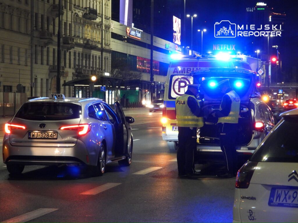 Policjanci w odblaskowych kamizelkach rozmawiają obok radiowozu na oświetlonej ulicy, obok zatrzymanego taksówki.