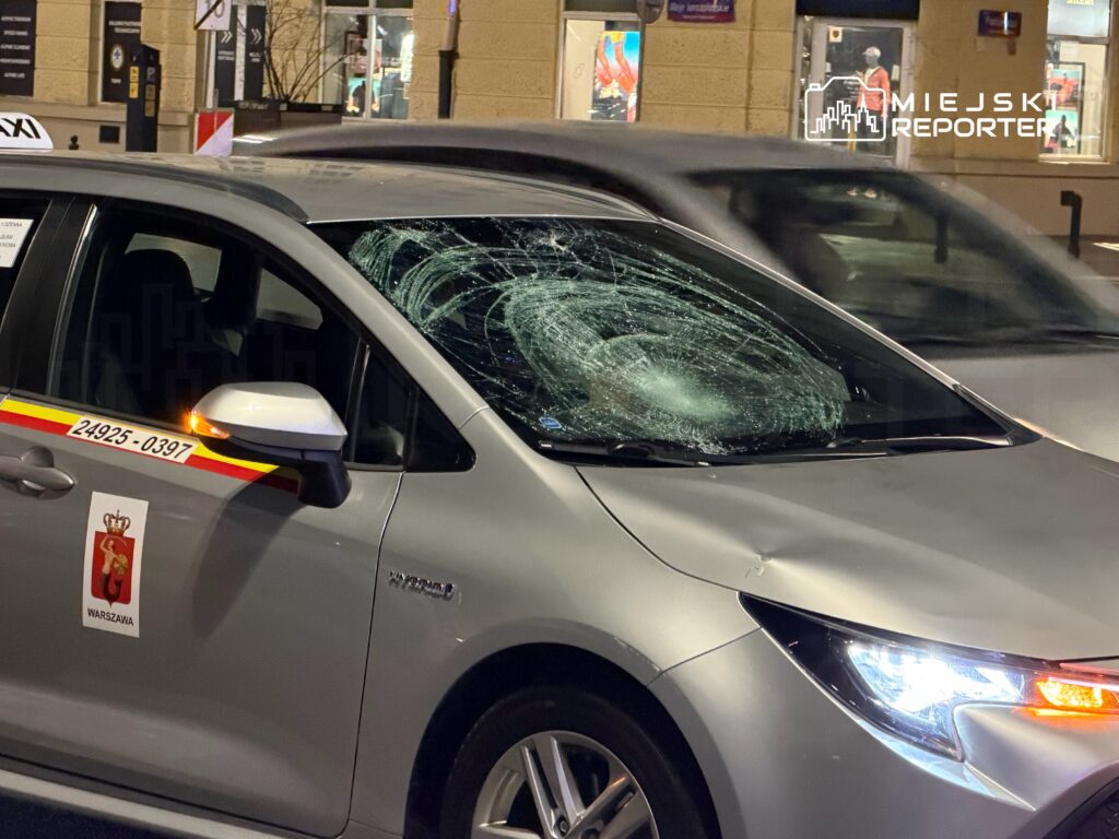 Zniszczona szyba przednia taksówki z widocznymi śladami uderzenia w nocy na miejskiej ulicy.
