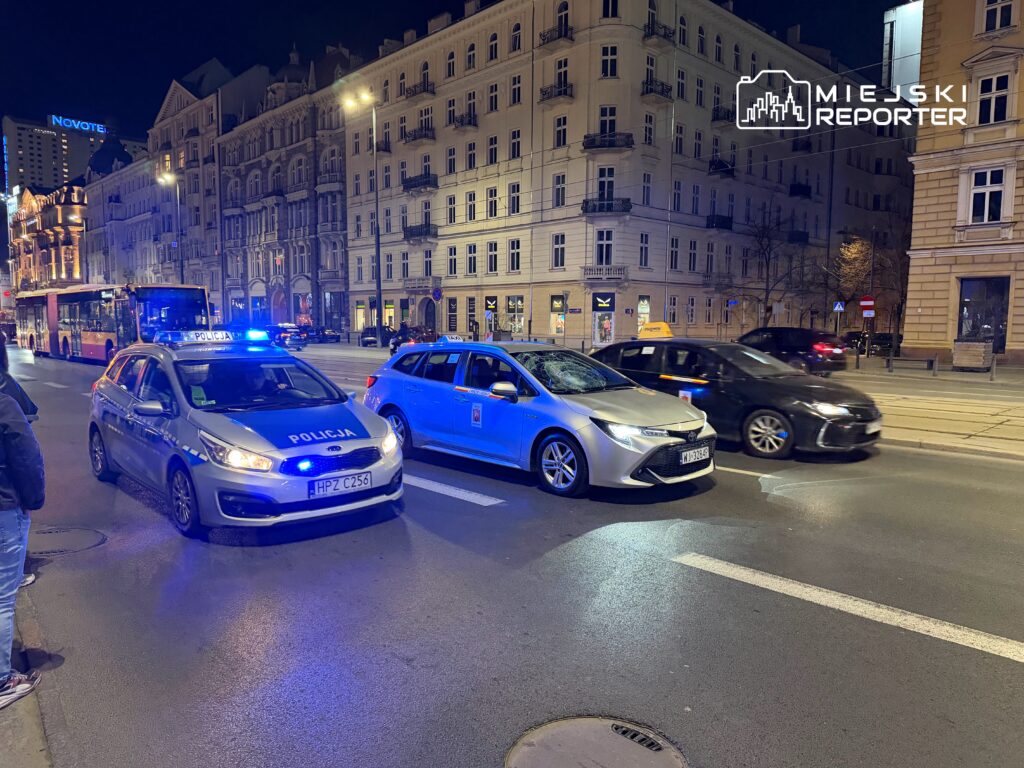 Policyjny radiowóz z włączonymi sygnałami świetlnymi zatrzymany na ulicy, obok przejeżdżające samochody w nocy.