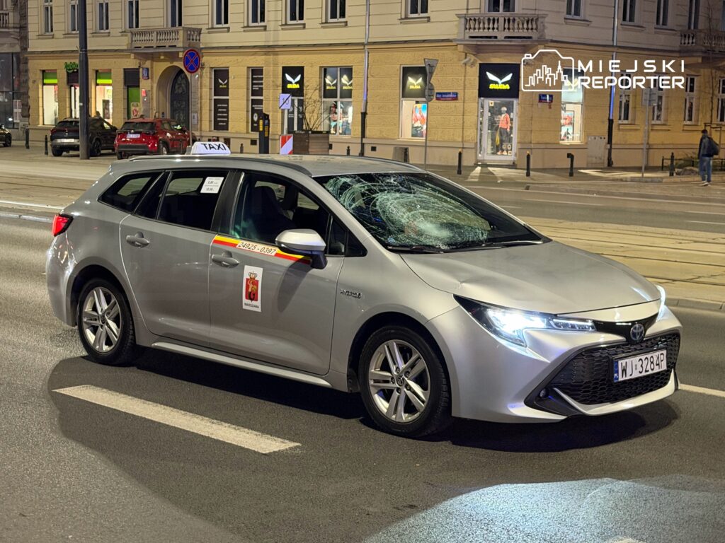 Uszkodzone taksówka z pękniętą szybą przednią zaparkowana na ulicy w nocy, w tle widoczne sklepy.