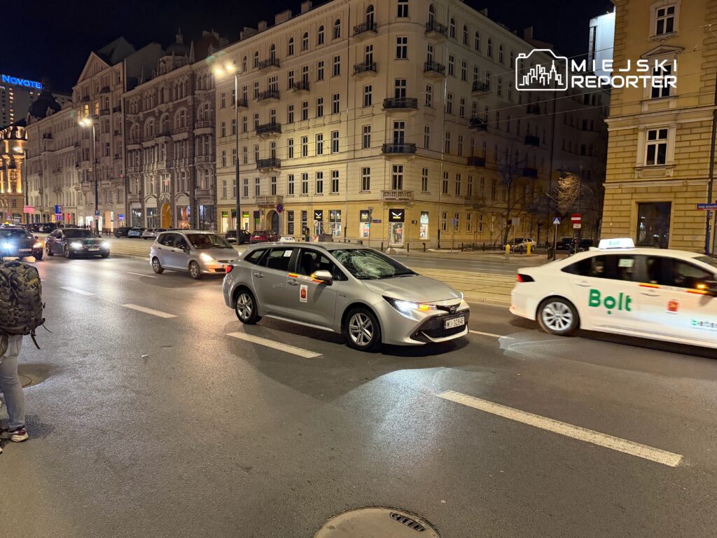 Samochody osobowe poruszają się po oświetlonej ulicy, obok zabytkowych budynków w nocy. Widać taksówki oraz pojazdy z logiem Bolt.