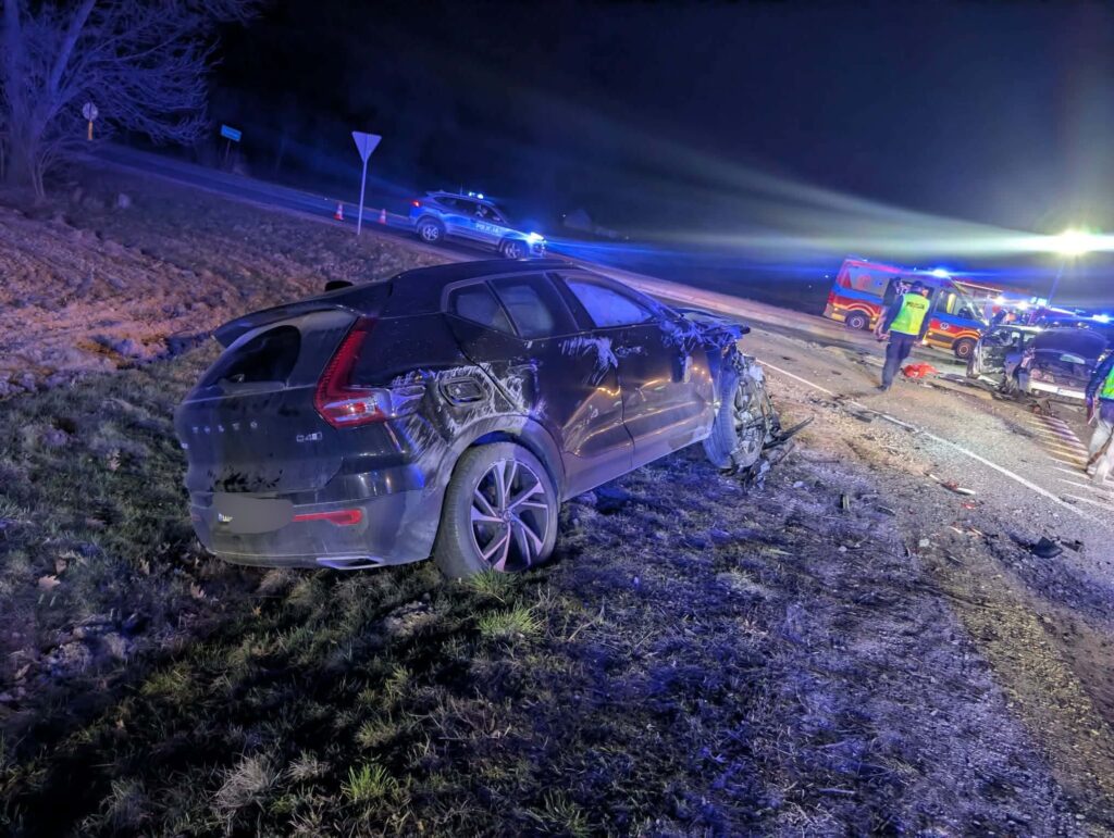 Uszkodzony czarny samochód stoi na poboczu drogi, obok interweniujący policjanci i zespół ratunkowy.