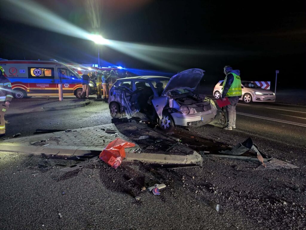 W nocy, na drodze, strażacy oraz ratownicy medyczni pracują przy rozbitym samochodzie osobowym.