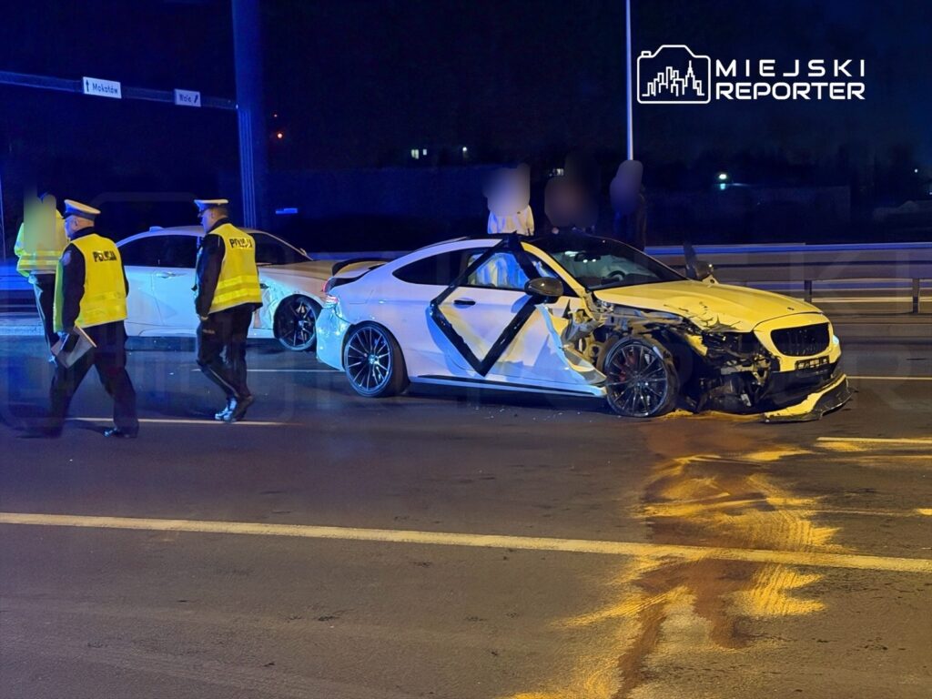 Dwaj policjanci w żółtych kamizelkach zabezpieczają miejsce wypadku drogowego, gdzie uszkodzone są dwa samochody osobowe.