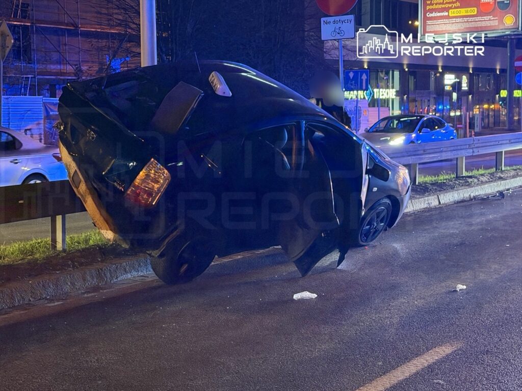 Czarny samochód osobowy unosi się na boku przy krawężniku, obok drogi, w otoczeniu miejskiego oświetlenia.