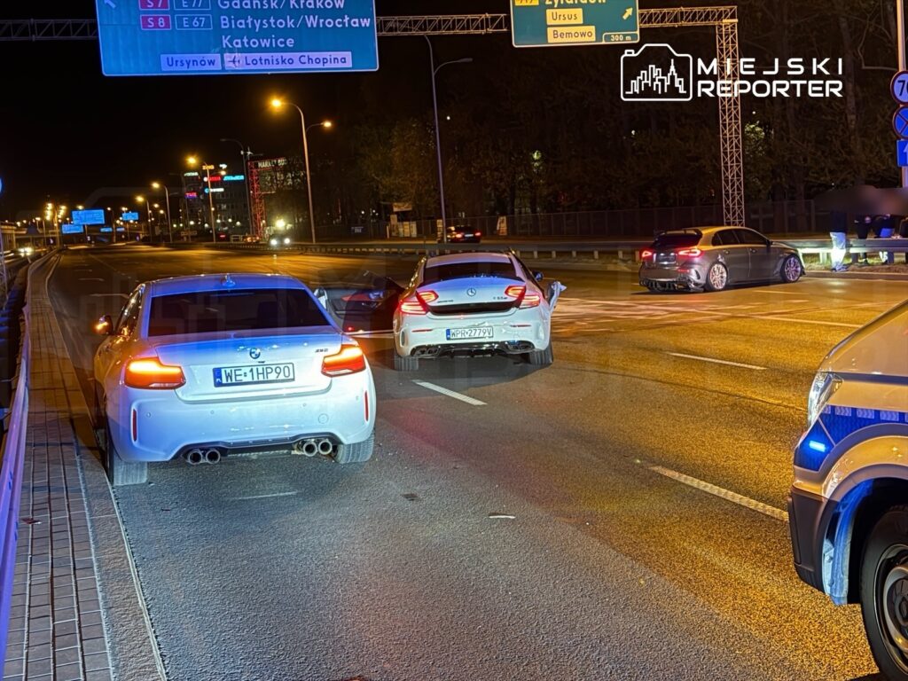 Dwa samochody osobowe zatrzymane na pasie ruchu, w tle znaki drogowe i oświetlone miasto.