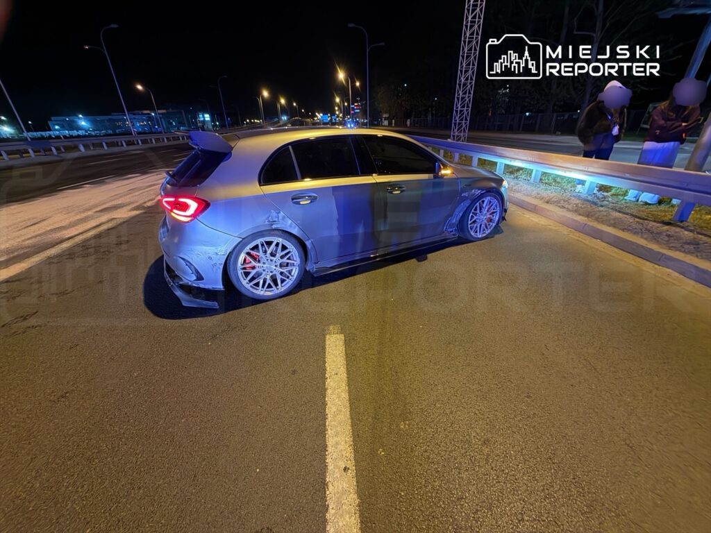 Samochód sportowy z przeszkloną karoserią zaparkowany na nocnej ulicy, obok grupka osób rozmawiających.