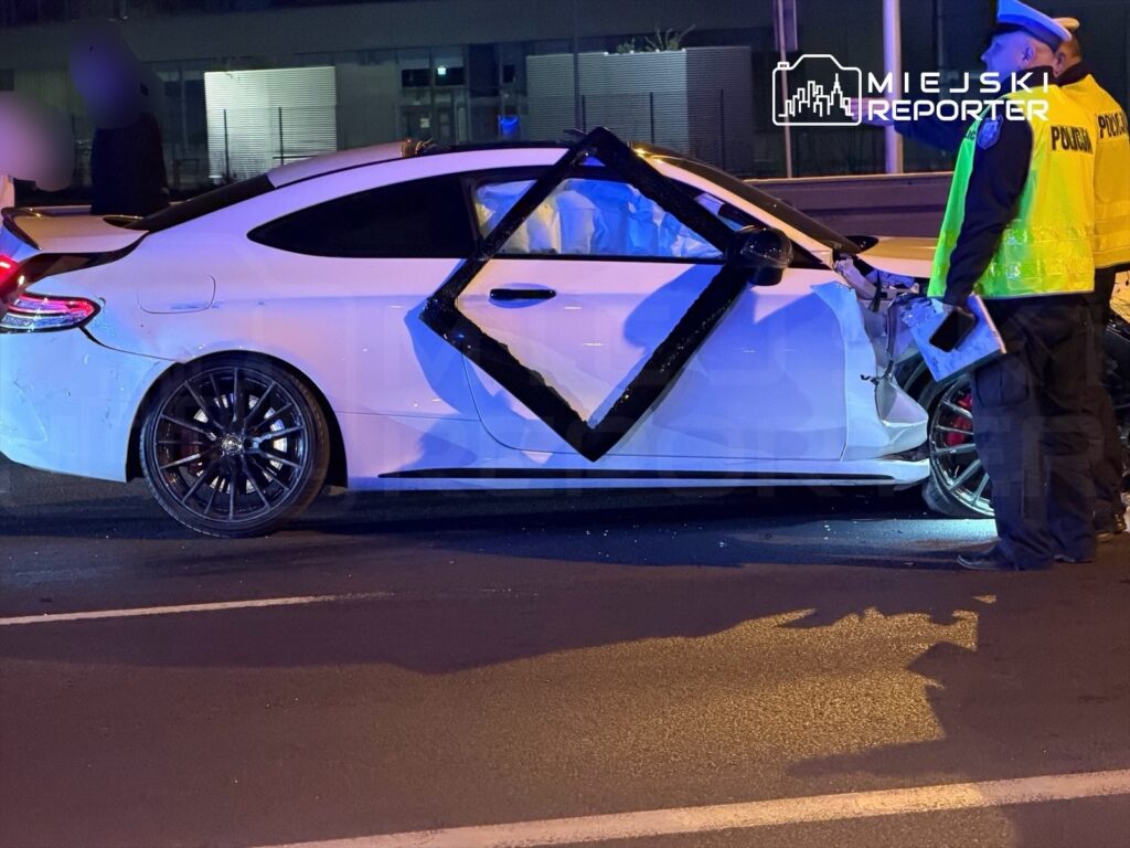 Policjant w odblaskowej kamizelce bada uszkodzone białe auto sportowe na drodze w nocy.