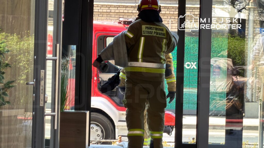 Strażak w uniformie z wężem gaśniczym wchodzi do budynku, w tle widoczny czerwony wóz strażacki.