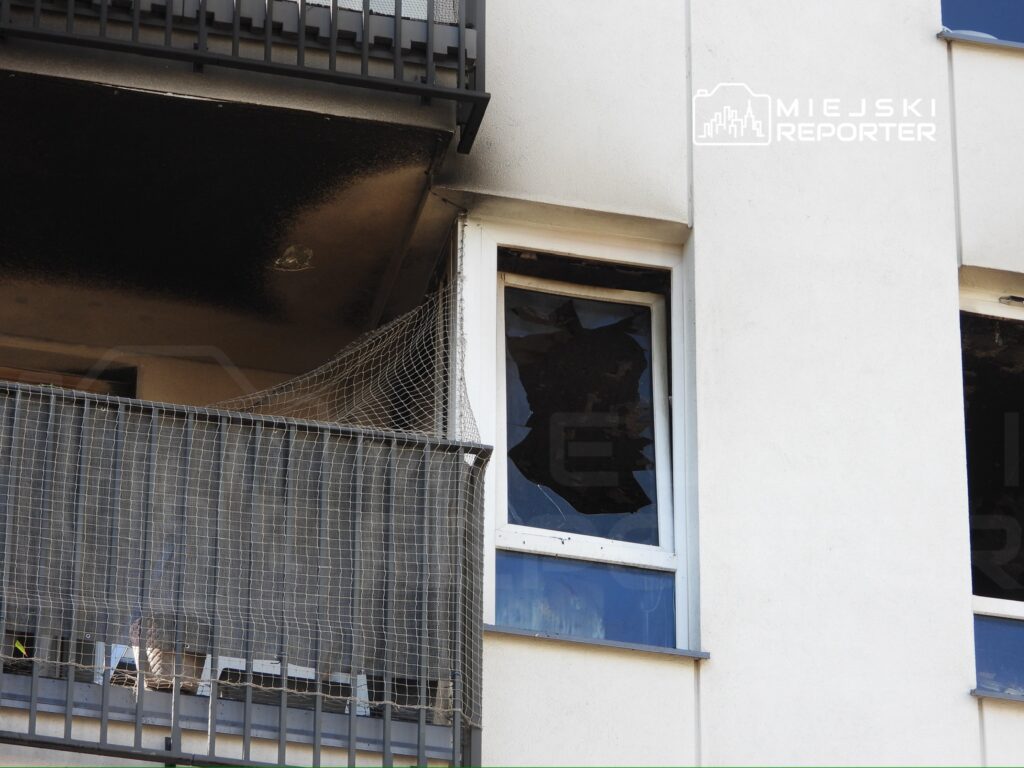 Zniszczona elewacja budynku z uszkodzonymi oknami i popiołem na balkonie.