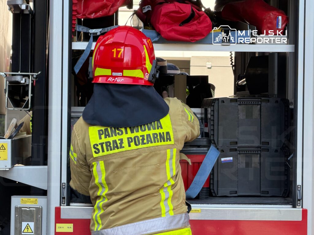 Strażak w hełmie z numerem 17 wyciąga sprzęt z wnętrza wozu strażackiego, przygotowując się do akcji.