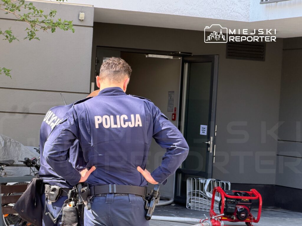 Dwaj policjanci w mundurach stoją przed otwartymi drzwiami budynku, rozmawiając ze sobą.