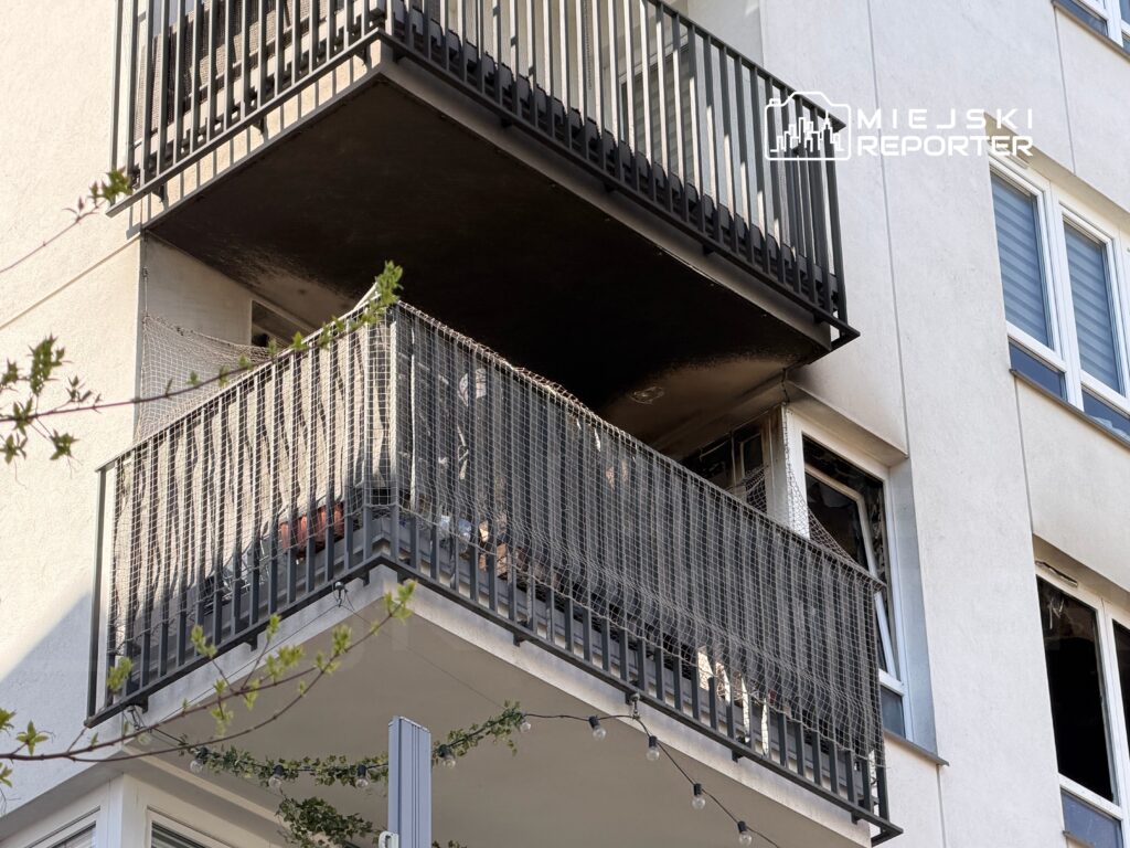 Czarny balkon z uszkodzeniami, na którym widoczne są zniszczone rośliny oraz siatka ochronna, w nowoczesnym budynku.