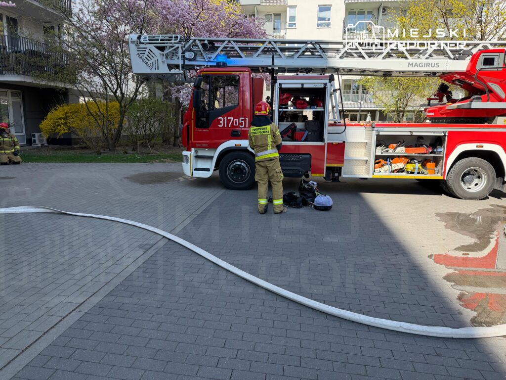 Strażak w mundurze przygotowuje sprzęt obok wozu strażackiego na osiedlowym podjeździe.