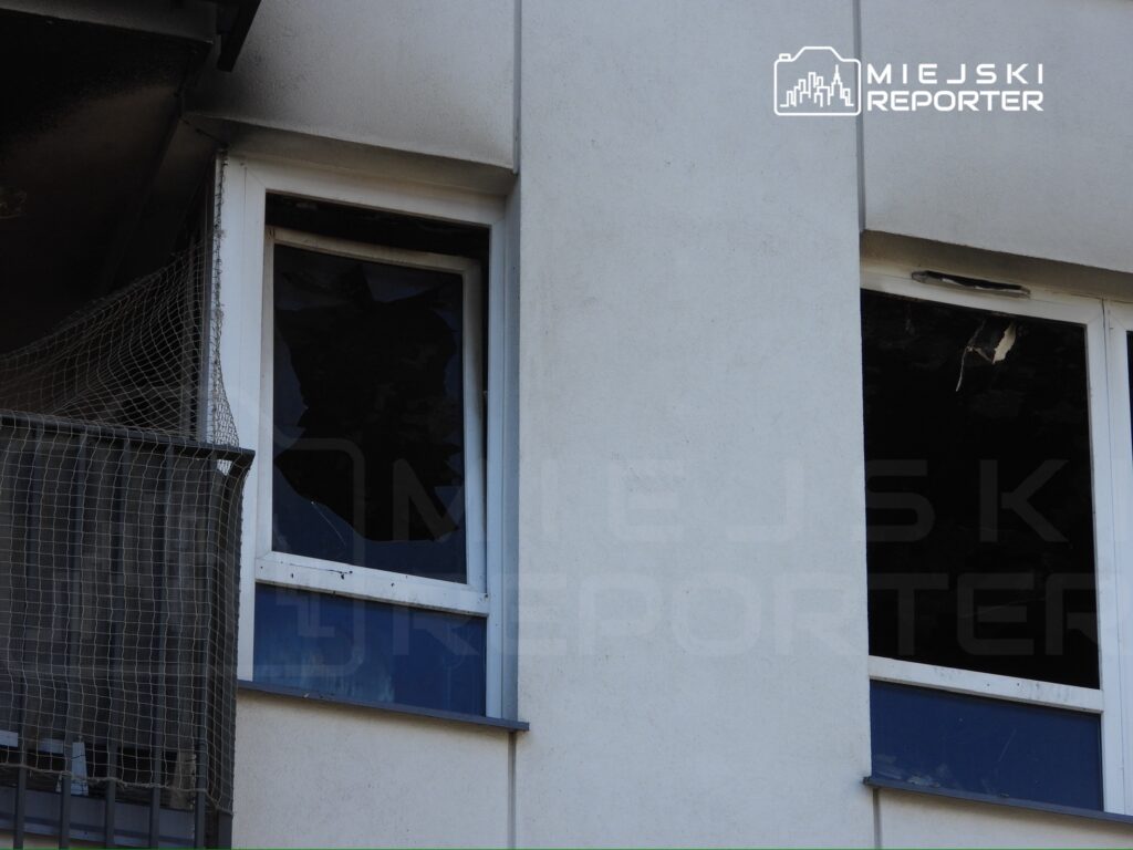Uszkodzone okna budynku z widocznymi pęknięciami i odłamkami szkła, z siatką ochronną na balkonie.