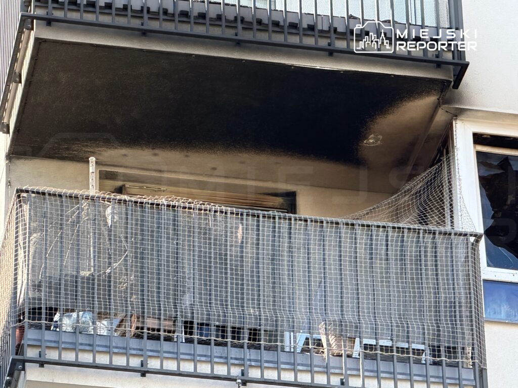 Czarny ślad po pożarze na balkonie mieszkania, z widocznym uszkodzeniem elewacji i zniszczonymi przedmiotami.