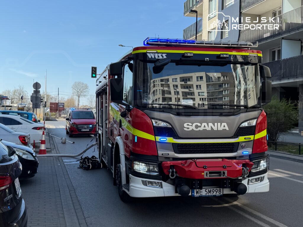 Czerwony wóz strażacki z włączonymi sygnałami świetlnymi stoi na ulicy, obok zaparkowanych samochodów.