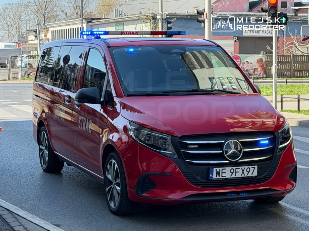 Czerwony samochód straży pożarnej z niebieskimi światłami zatrzymany na skrzyżowaniu.