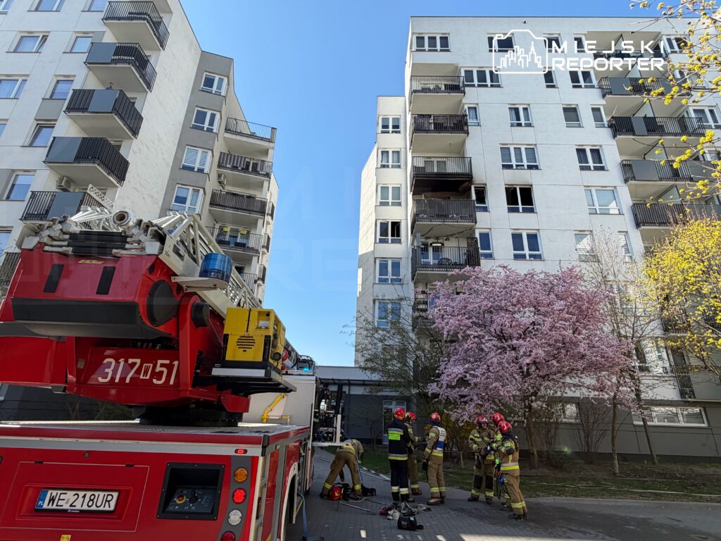 Strażacy w hełmach omawiają akcję ratunkową przed nowoczesnym budynkiem mieszkalnym z balkonami.