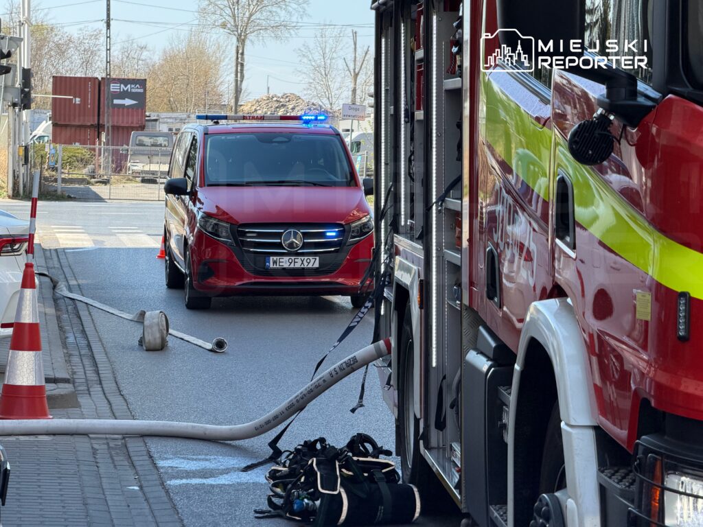 Czerwony pojazd ratunkowy z niebieskimi światłami zatrzymany przy drodze, obok wozu strażackiego i sprzętu gaśniczego.