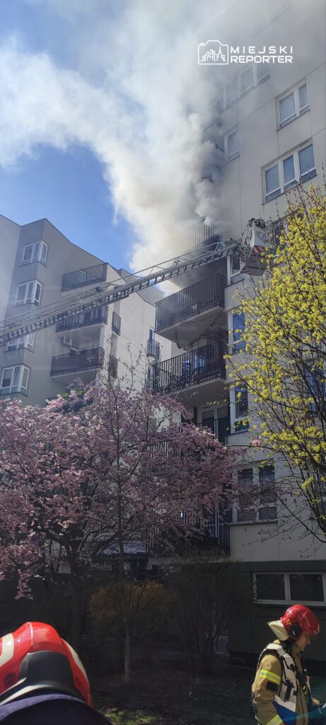 Gęsty dym wydobywający się z balkonu budynku, obok strażak w hełmie i mundurze, w tle kwitnące drzewa.