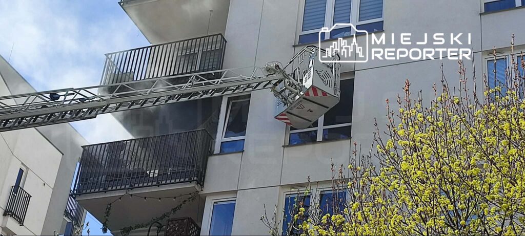 Strażak na podnośniku gaśniczym przystępuje do akcji ratunkowej przy balkonie budynku, z którego wydobywa się dym.