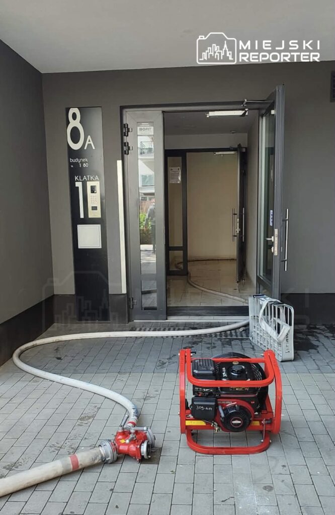 Woda wypływa z budynku, a czerwony pompa stoi obok węża przy drzwiach wejściowych.