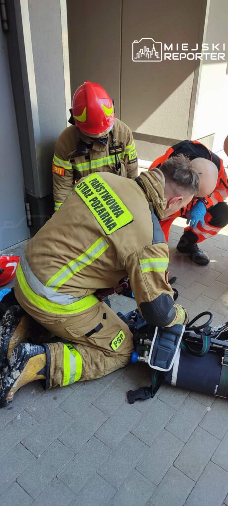 Grupa strażaków w ubraniach ochronnych prowadzi działania ratunkowe na chodniku, obsługując sprzęt gaśniczy.