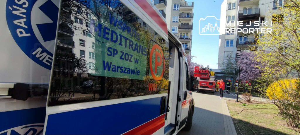 Karetka pogotowia z widocznym napisem "MEDITRANS SP ZOZ w Warszawie" stoi na osiedlowej drodze, w tle strażacy w akcji.