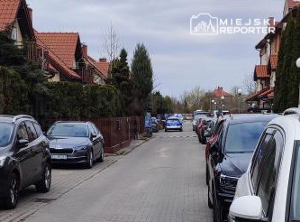 Ulica z rzędem zaparkowanych samochodów, w tle widać domy jednorodzinne z ogródkami.