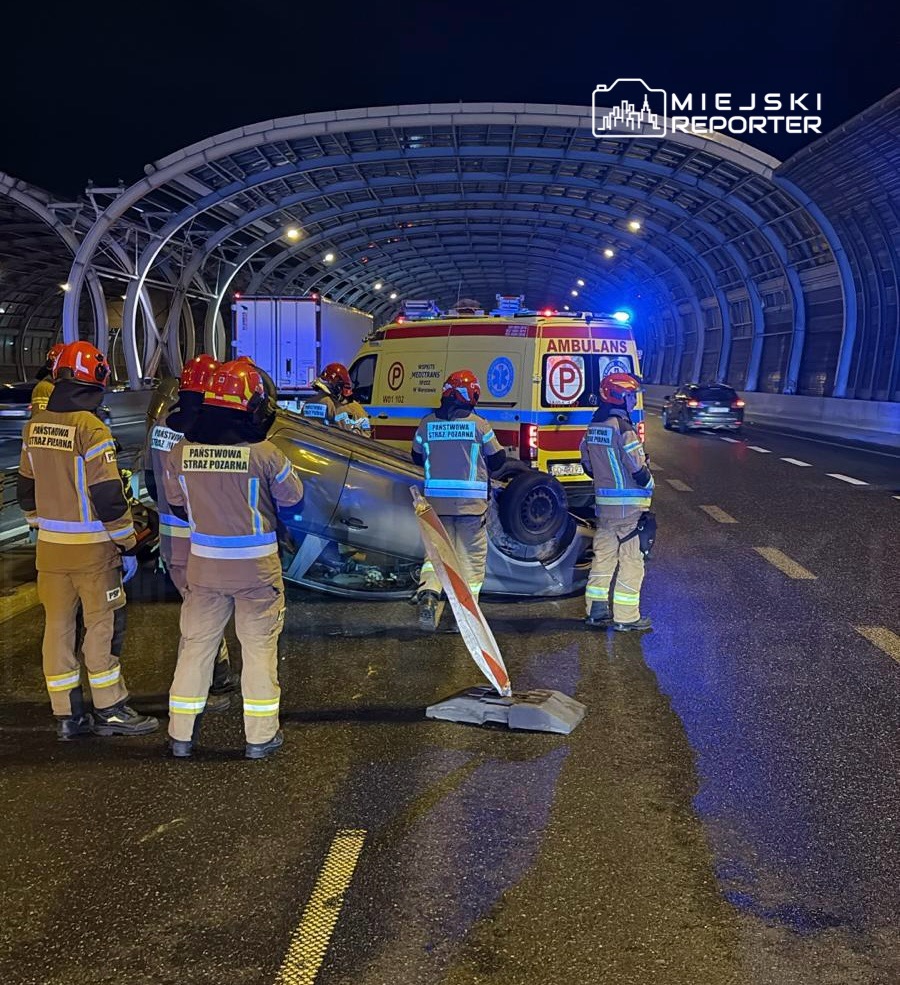 Grupa strażaków w mundurach zabezpiecza miejsce wypadku drogowego, gdzie przewrócone auto leży na boku.