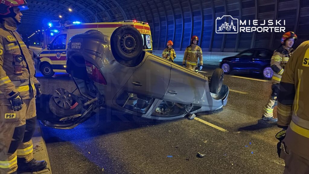 Strażacy w mundurach pracują przy przewróconym samochodzie w tunelu drogowym, w tle widać karetkę.