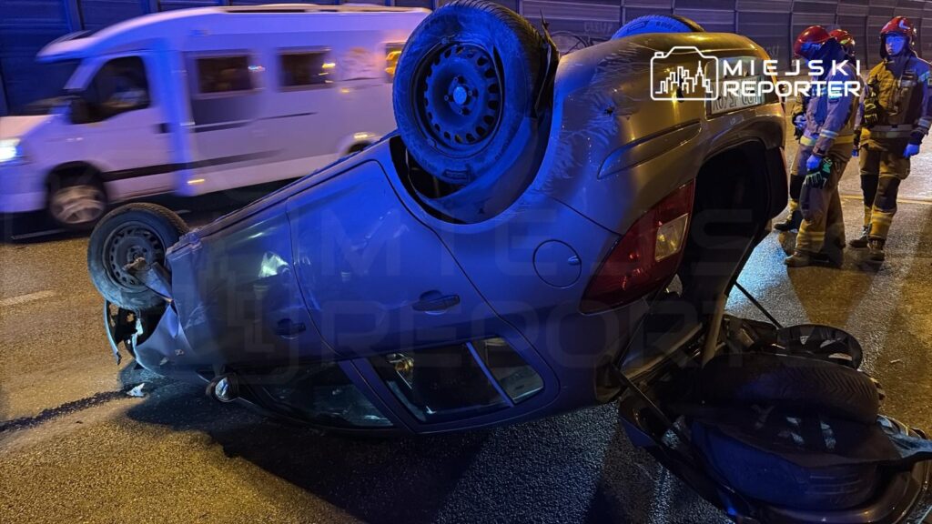 Przewrócony samochód leży na boku na jezdni, obok grupy strażaków w hełmach.