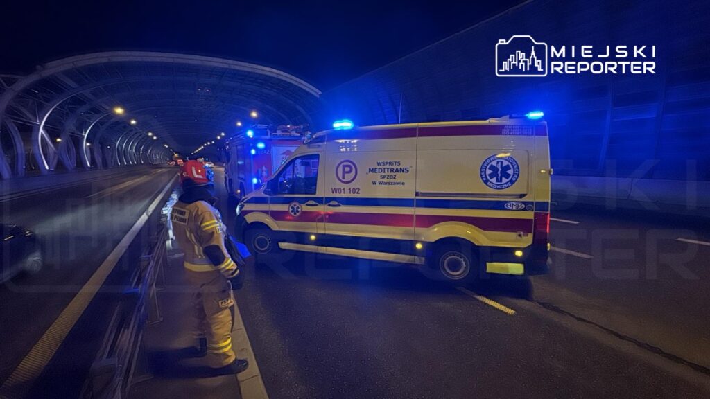 Ratownik w żółtym mundurze obserwujący ambulans z włączonymi sygnałami świetlnymi na oświetlonym tunelu.