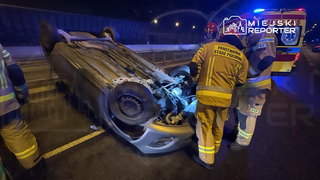 Strażacy w mundurach interweniują przy przewróconym samochodzie na drodze w nocy, obok widać ambulans.