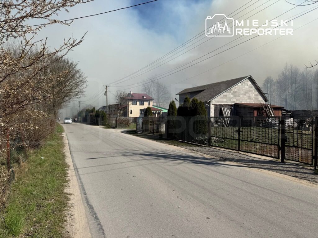 Dym unoszący się nad zabudowaniami mieszkalnymi przy drodze, w tle widoczny dom z czerwonym dachem.