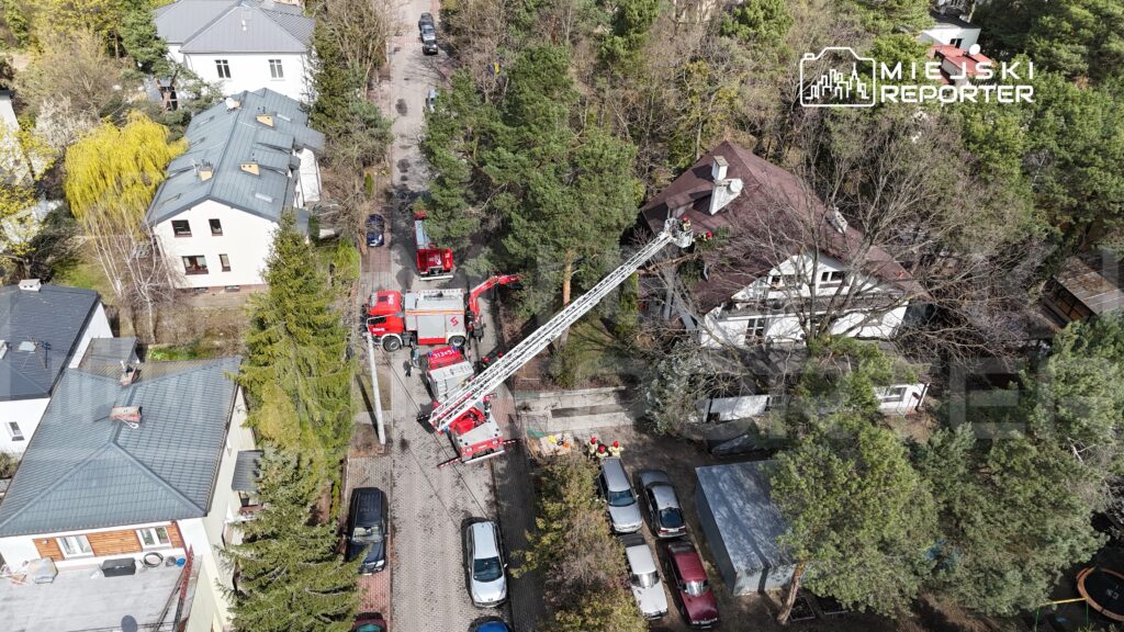 Strażacy na drabinie wykonują prace ratunkowe na dachu budynku mieszkalnego w otoczeniu drzew i zaparkowanych samochodów.