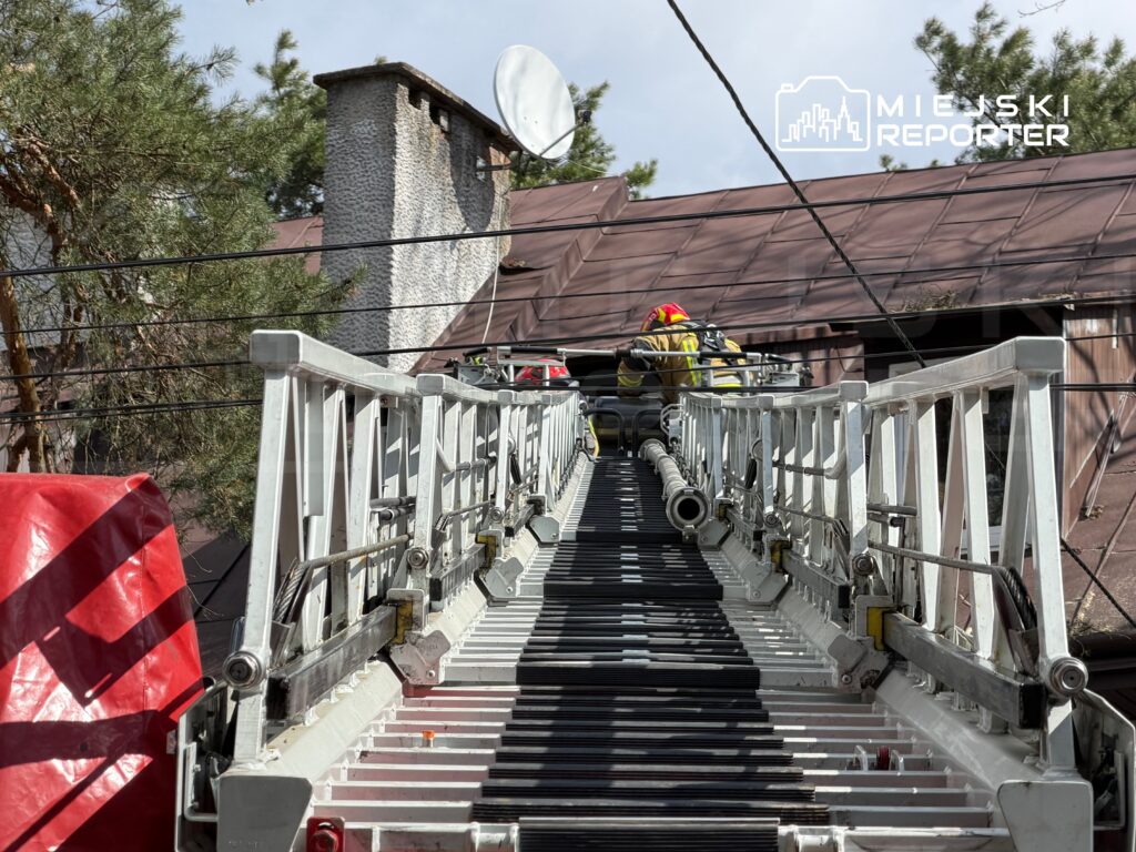 Strażak w kasku i mundurze wspina się po drabinie strażackiej w kierunku dachu budynku.