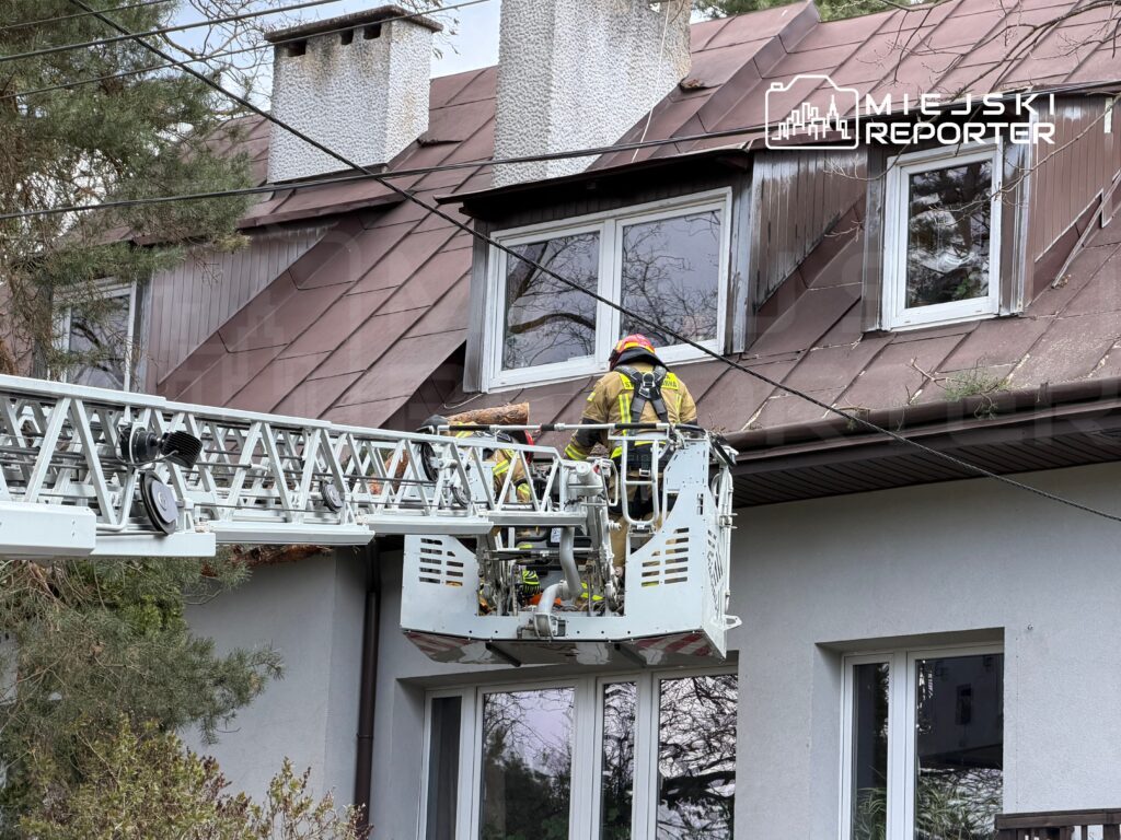 Strażak w hełmie pracuje na platformie podnośnika przy dachu domu, sprawdzając okna.
