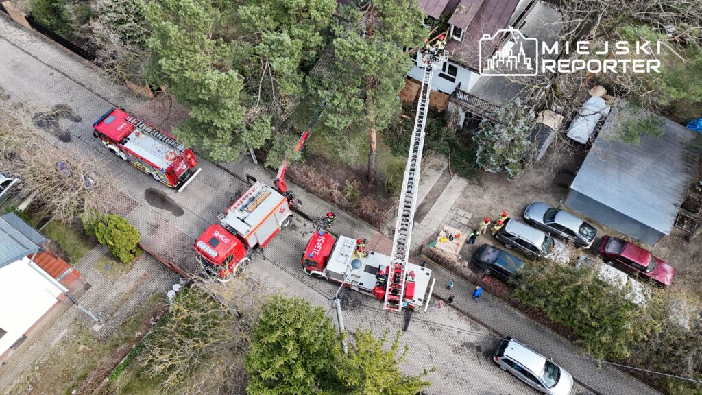 Strażacy pracują przy akcji ratunkowej, używając drabiny i wozów strażackich na osiedlowej ulicy.