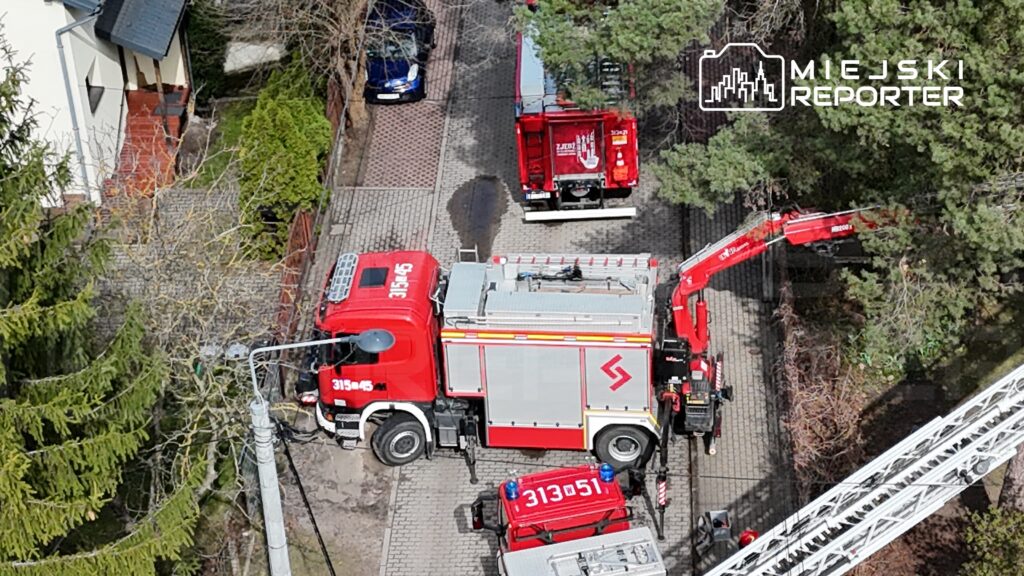 Czerwony wóz strażacki z podnośnikiem oraz inne pojazdy strażackie zaparkowane na wąskiej ulicy w otoczeniu drzew.