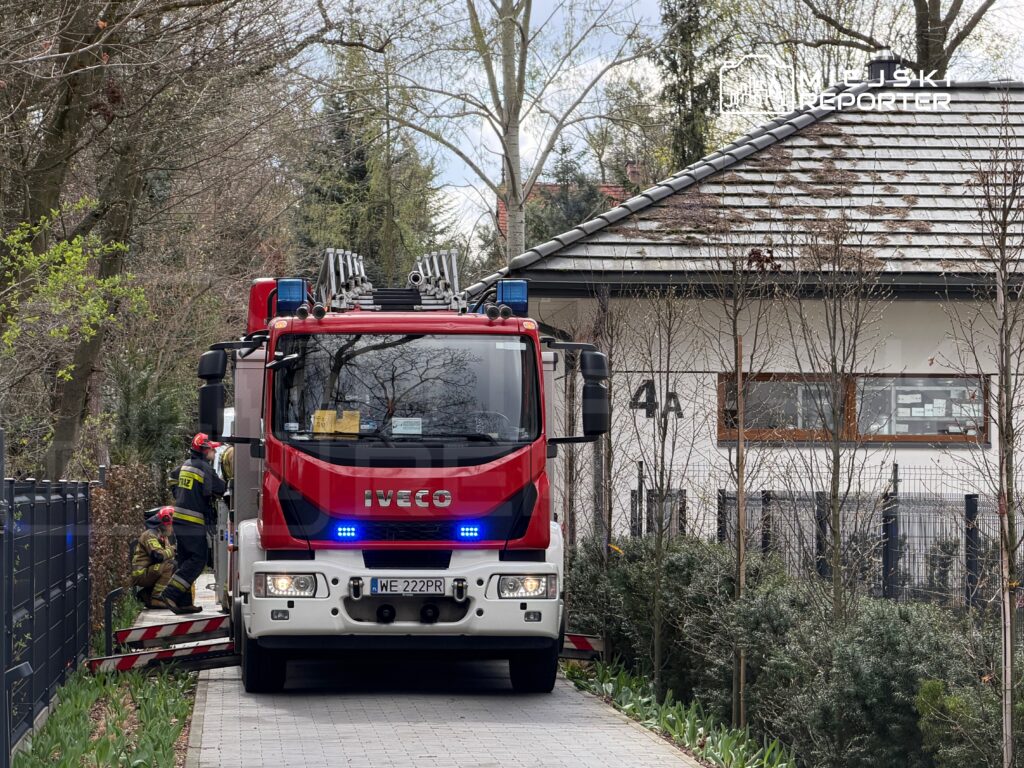 Czerwony wóz strażacki z włączonymi sygnałami świetlnymi zatrzymany przed budynkiem mieszkalnym, obok strażacy w mundurach.