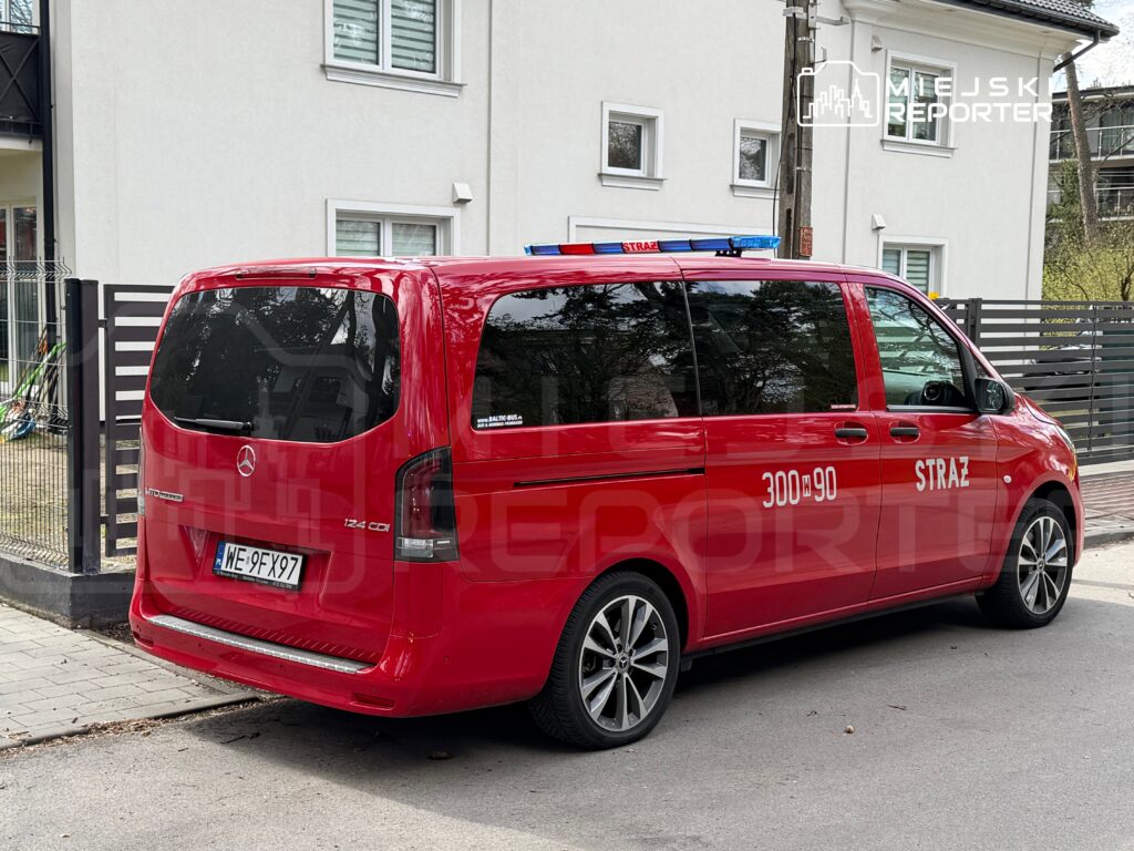 Czerwony samochód strażacki zaparkowany przed budynkiem mieszkalnym, z widocznymi oznaczeniami i numerem alarmowym.