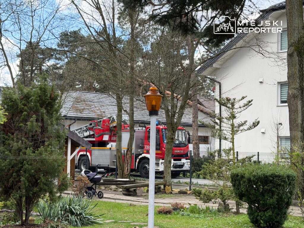 W tle widoczna strażacka ciężarówka z wysięgnikiem, zaparkowana obok nowoczesnego budynku mieszkalnego. W pierwszym planie wózek dziecięcy na trawniku.