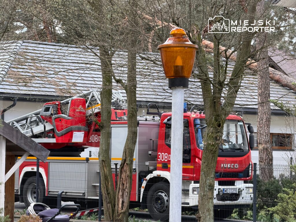 Czerwony wóz strażacki z drabiną parkowany obok domu, otoczony przez drzewa w miejskim otoczeniu.