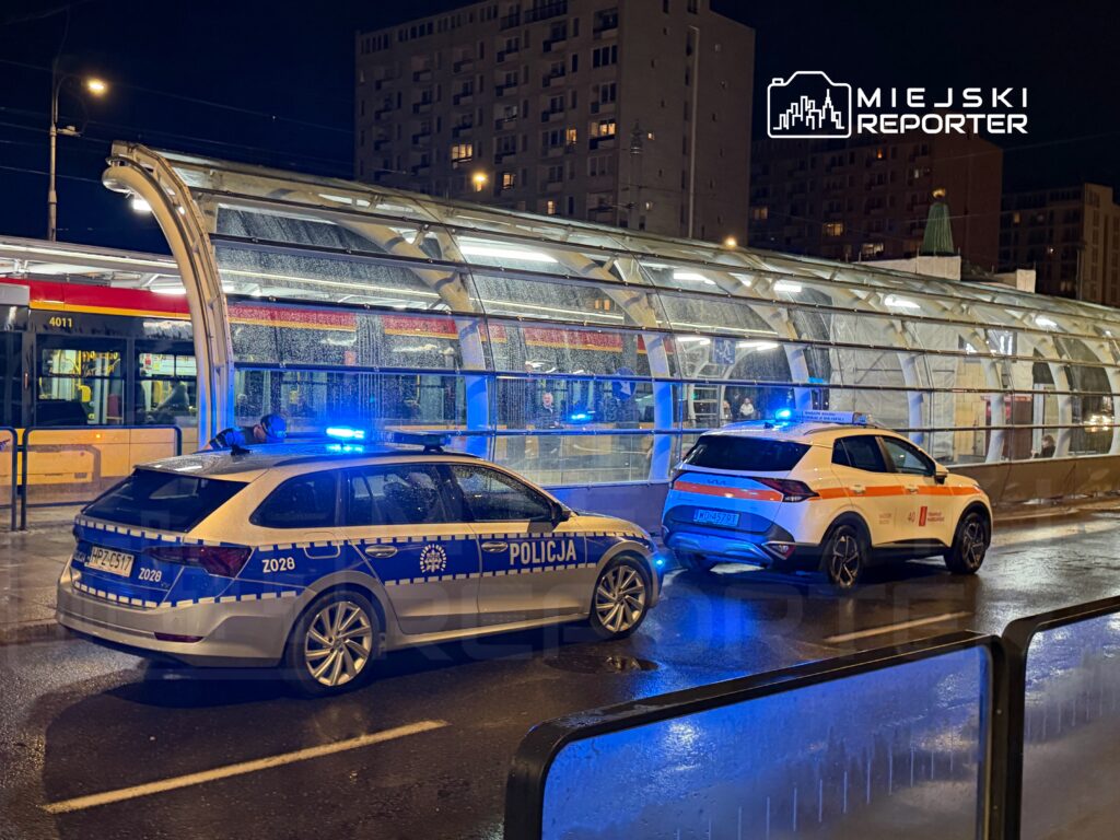 Policyjny radiowóz z niebieskimi światłami stoi obok pojazdu służb ratunkowych na oświetlonym przystanku w deszczową noc.