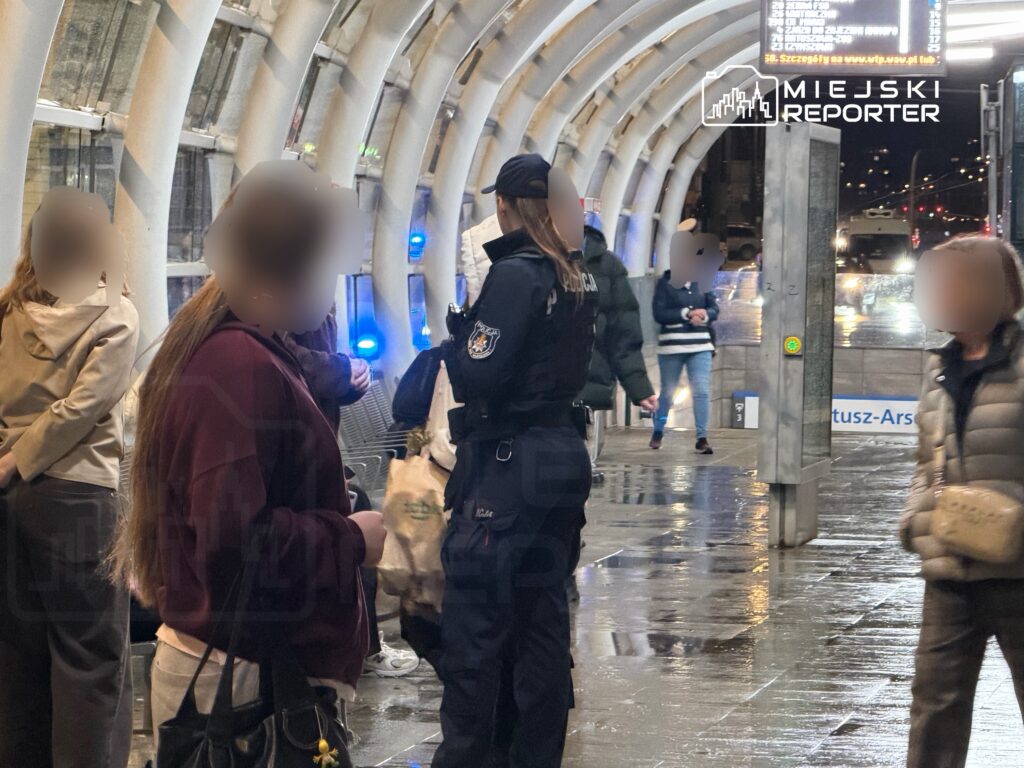 Policjant w mundurze rozmawia z grupą osób na przystanku, w tle wyświetlacz z informacjami o odjazdach.